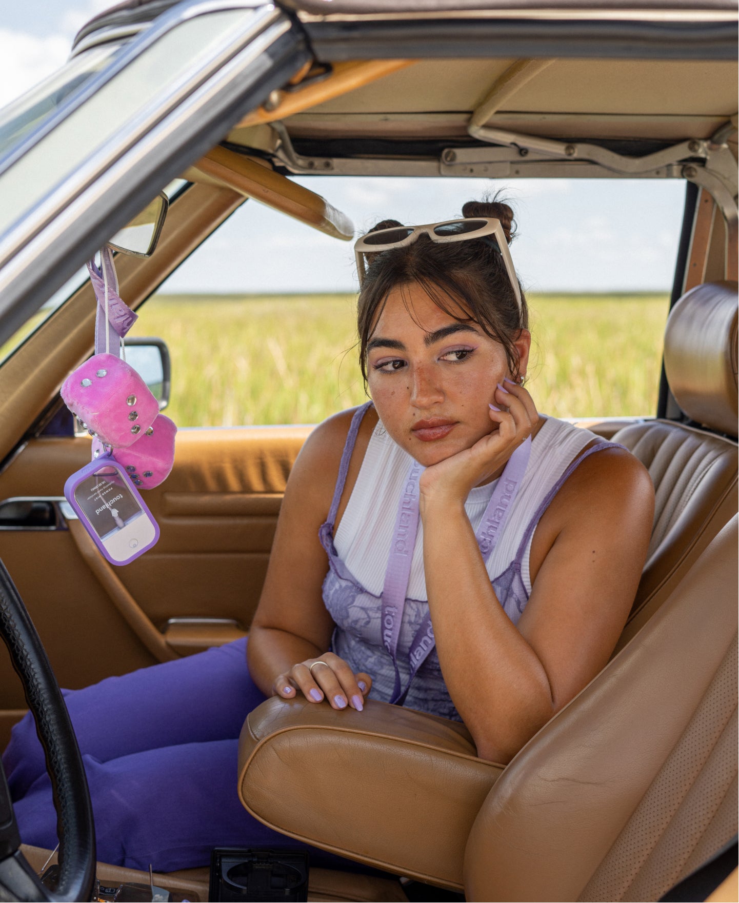 Female model is resting her arm on the middle arm rest of a vintage car, on the rearview mirror a lilac lanyard with a power mist bottle is hanging down