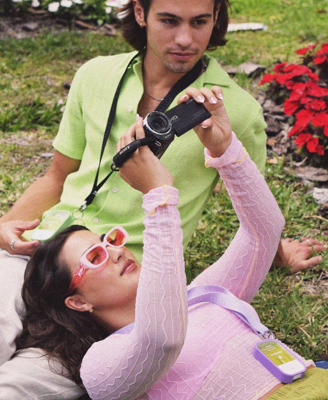 A girl and a boy laying in the grass looking at film wearing Touchland's Aloe You & Pure Lavender Power with matching lanyard and case.