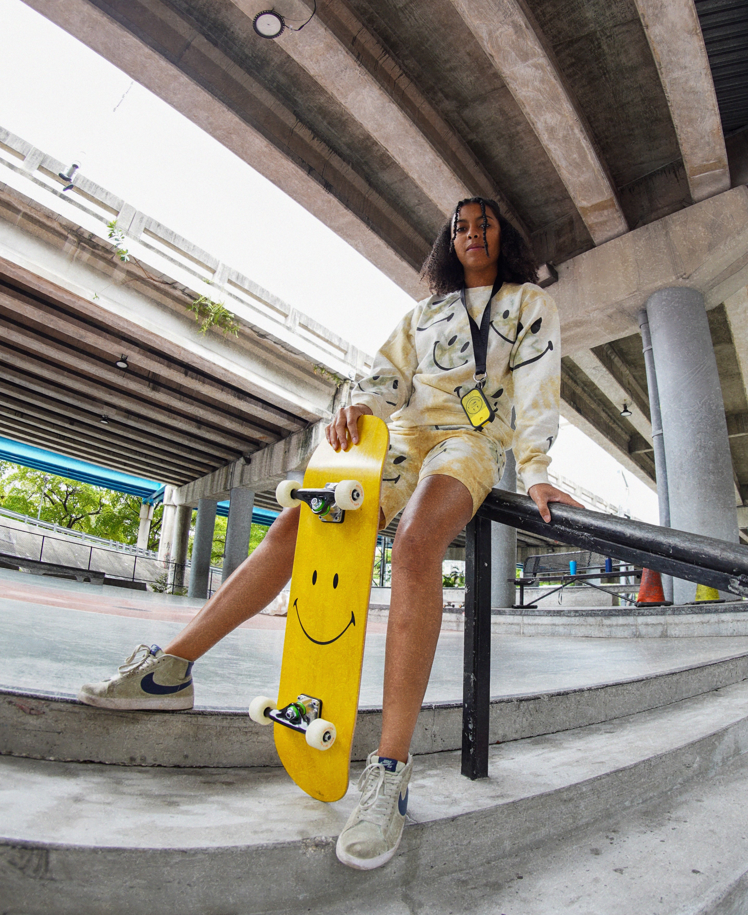 Female model resting on a skatepark rail, holding a yellow Smiley skateboard with a Touchland Black Lanyard with a Power mist hanging