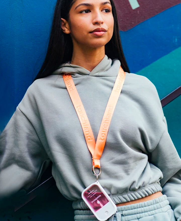 Girl in grey matching sweat set with dark hair wearing orange lanyard with white case and berry pink mist around her neck and looking off camera